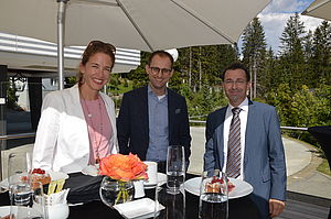 Eine Gruppe von drei Personen steht im Freien unter einem Sonnenschirm und lächelt. Sie stehen neben einem Tisch mit Essen, Getränken und einer Vase mit orangefarbenen Rosen vor einer Kulisse aus Bäumen und blauem Himmel.