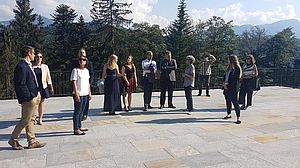 Eine Gruppe von Menschen steht auf einer sonnenbeschienenen Terrasse mit Bergen und Bäumen im Hintergrund. Sie scheinen sich zu unterhalten und die schöne Aussicht zu genießen.