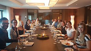 Eine Gruppe von 14 Personen sitzt um einen langen Esstisch in einem eleganten Raum. Sie lächeln, vor ihnen stehen Teller mit Essen und Gläser mit Wein.
