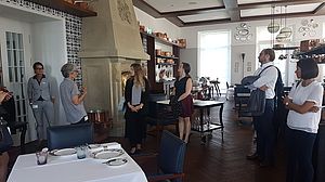 In einem hellen, eleganten Speisesaal mit Holzfußboden und großen Fenstern sitzt eine Gruppe von Menschen beiläufig zusammen. Die Atmosphäre ist entspannt und gesprächig.