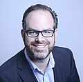 Ein Mann mit Bart und Brille lächelt warmherzig, er trägt eine Anzugsjacke und ein weißes Hemd vor einem hellblauen Hintergrund. Professioneller und sympathischer Ton.