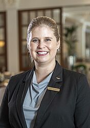 Eine Person in einem dunklen Blazer lächelt freundlich in die Kamera. Sie steht in einem eleganten Essbereich mit eleganten Holzwänden und bequemen Sitzgelegenheiten.