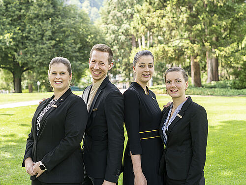 Vier Personen in schwarzen Uniformen stehen lächelnd nebeneinander in einem sonnigen Park mit grünem Gras und hohen Bäumen und vermitteln eine professionelle und doch entspannte Stimmung.