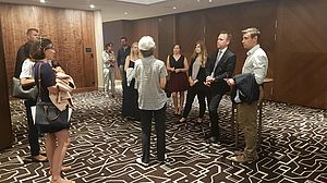 Eine Gruppe von Personen steht in einer Hotellobby mit einem geometrisch gemusterten Teppich und Holzwänden. Sie scheinen in eine Diskussion verwickelt zu sein und sind formell gekleidet.