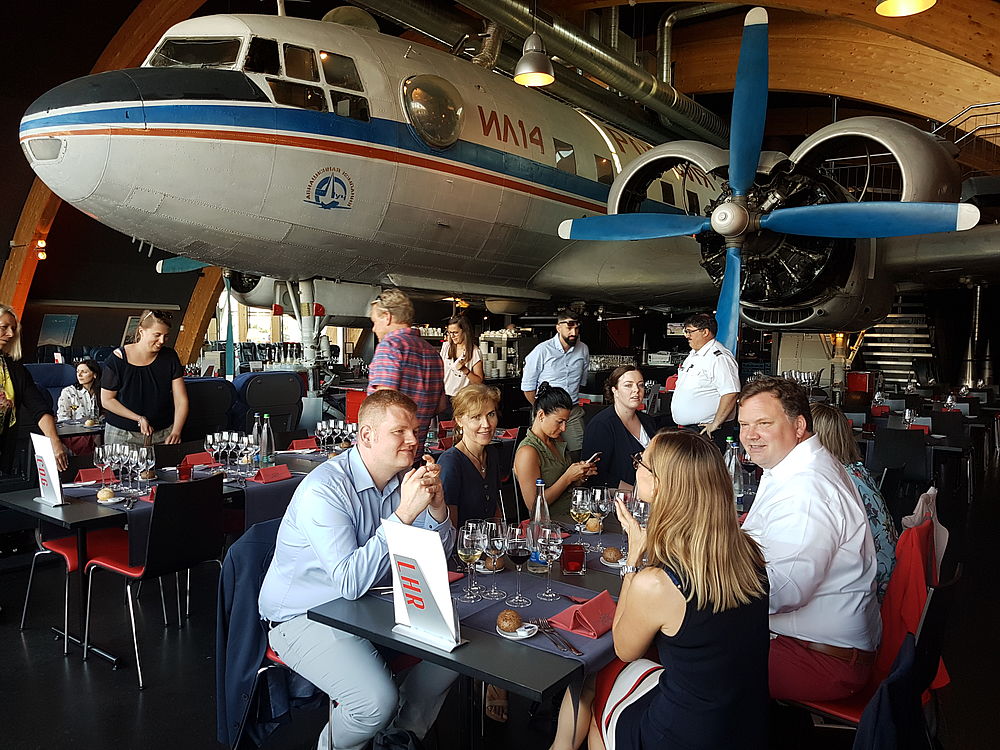 Die Gäste speisen unter einer großen Flugzeugausstellung in einer Museumsumgebung und genießen die Gespräche und das Essen. Die Stimmung ist lebhaft und gesellig, das Thema ist die Luftfahrt.