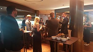 Eine Gruppe von formell gekleideten Personen unterhält sich bei einem schwach beleuchteten Empfang. Auf den Tischen stehen Snacks und Getränke und schaffen eine warme, gesellige Atmosphäre.