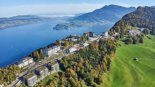 Luftaufnahme eines Luxushotels am See, umgeben von herbstlichen Bäumen und Bergen unter einem klaren blauen Himmel. Die friedliche Szene strahlt Ruhe und Schönheit aus.