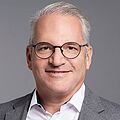 Ein lächelnder Mann mit grauen Haaren und Brille, der einen grauen Anzug mit weissem Hemd trägt. Er vermittelt Professionalität.