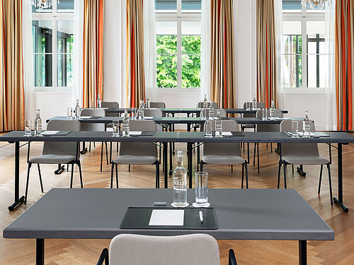 Ein geräumiger, eleganter Konferenzraum mit ordentlich angeordneten Tischen und Stühlen, großen Fenstern mit Vorhängen, Holzfußboden und üppigem Grün im Außenbereich, der einen professionellen Eindruck vermittelt.