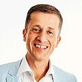 Eine freundlich lächelnde Person in einem hellblauen Blazer und weißem Hemd vor einem schlichten weißen Hintergrund. Die Stimmung ist freundlich und ansprechbar.