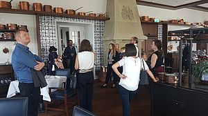 Eine Gruppe von Menschen steht in einem Restaurant mit eleganter Einrichtung, Kupfertöpfen auf Regalen und blau gekachelten Wänden. Die Stimmung ist locker und gesellig.