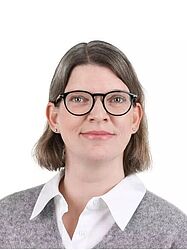 Eine Frau mit schulterlangem braunem Haar und Brille lächelt leicht. Sie trägt einen grauen Pullover über einem weißen Hemd mit Kragen vor einem schlichten Hintergrund.