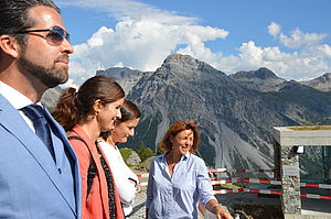 Vier Menschen lächeln und genießen einen sonnigen Tag auf einem Bergbalkon. Der strahlend blaue Himmel, die Wolken und die zerklüfteten Berggipfel schaffen eine lebendige und fröhliche Szene.